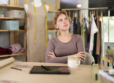 Young sad female dressmaker sitting at the table in sewing workshopの写真素材