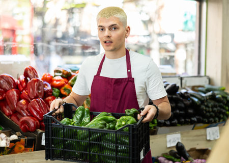 Man salesperson carries large basket of bell pepper from warehouse to sales area in shopの写真素材