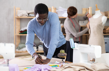 African man draw dress pattern of product on paper. Companion work in background with mannequinの写真素材