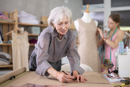 Mature tailor draws of a fashion design against the background of a assistant