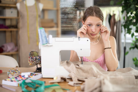 Tired young tailoress sitting at the sewing-machine in tailors shop