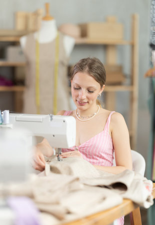 Positive young female dressmaker working with sewing-machine in tailors workshopの写真素材