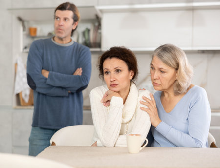 Man quarrels with his wife in kitchen, standing behind her, and elderly mother hugs and calms her daughterの写真素材