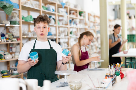 Teenage boy in an apron demonstrates clay mugs, which he himself painted with colored paintsの写真素材