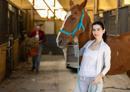 Female worker takes horse out of stall, holds animal by bridle and leads pet for walkの写真素材