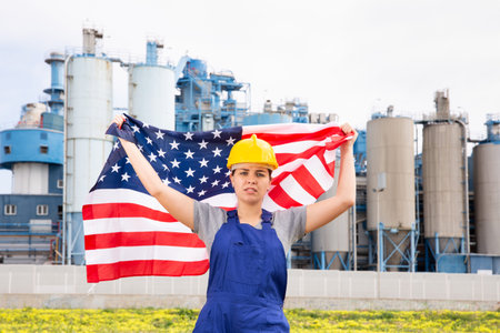 Worried girl in work clothes and hardhat with flag of usa standing in front of industrial scenery