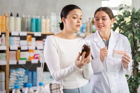 Asian woman buyer choosing medicinal cough liquid syrup standing between shelves at pharmacy, young girl pharmacist consulting customerの写真素材