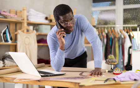 Tailor communicating on phone and reviewing specifications on laptop in workshopの写真素材