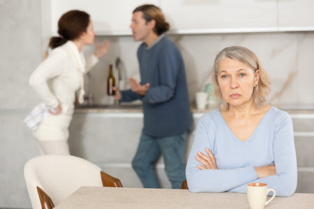 Frustrated senior woman is sitting in kitchen during quarrel between her son and his wifeの写真素材