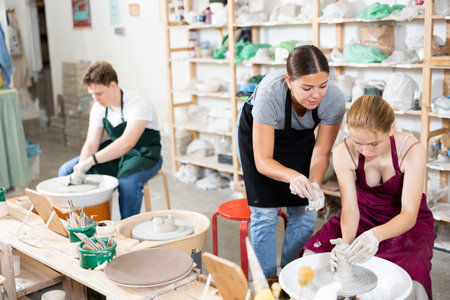 Young woman teacher helps teenagers boy and girl to make product on potters wheelの写真素材