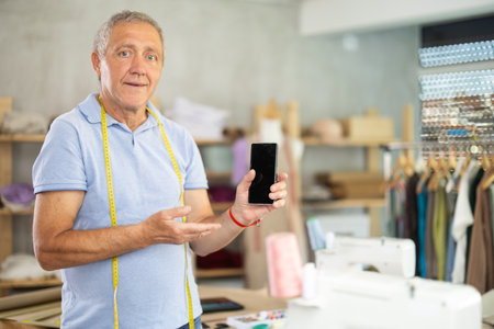 Senior man tailor in studio shows phone, points finger at touchscreen of smartphoneの写真素材