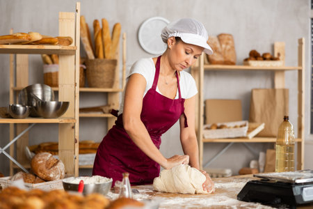 Skilled happy young woman baker in apron standing at work table, kneading dough for baking bread in...
