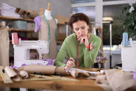 Thoughtful female tailor designs new clothes by drawing on white paper in workshopの写真素材