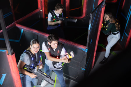 Laser tag players positive smiling guys and girls playing in teams in dark laser tag room. View from...