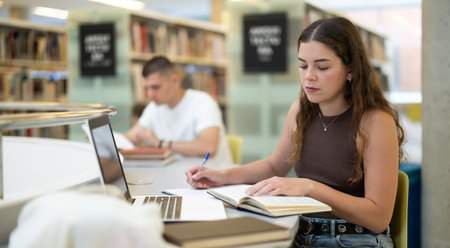 Portrait of positive woman with laptop and book in libraryの写真素材