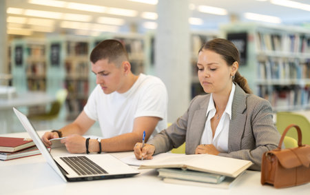 Girl and boy studying for exam together in library - using books and laptopの写真素材
