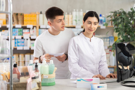 Male customer holding prescription searches for medicine in pharmacy. Female pharmacist offers to help find information on computerの写真素材