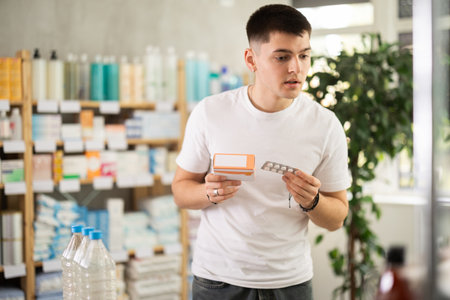 Portrait of handsome young guy holding and choosing box with best pills to improve well-being in pharmacyの写真素材