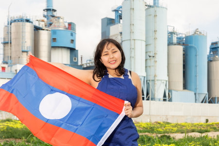 Portrait of teenage girl in blue overalls with flag of Laos in her hands against the backdrop of modern metallurgical plantの写真素材