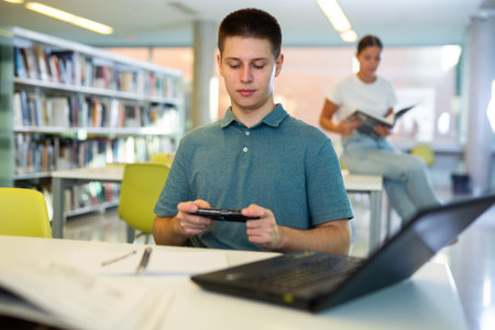 Young casual male student watching video on mobile phone after using laptop in the library
