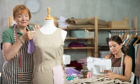 Elderly dressmaker stands near a mannequin and checks the dress against the background of the assistantの写真素材
