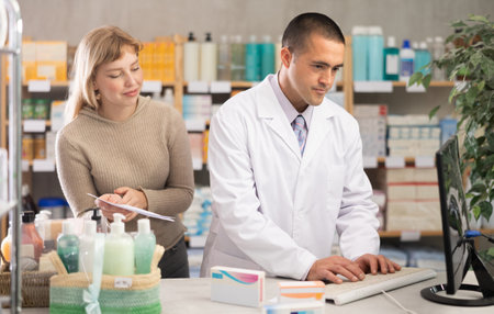 Young male pharmacist working in computer while female client standing with prescription behindの写真素材
