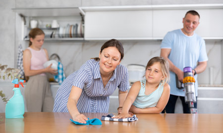 Cheerful woman wiping table in kitchen with preteen daughterの写真素材