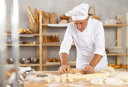 Senior male baker works with rolling pin, rolls out and spreads out pieces of dough
