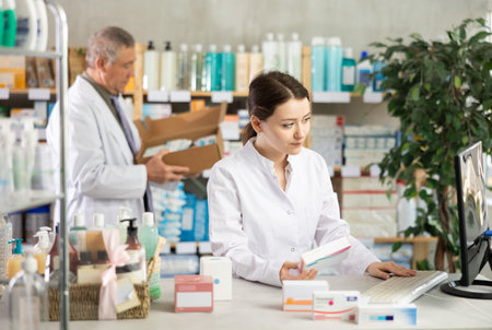 Girl pharmacist uses computer at workplace. Male colleague in backgroundの写真素材