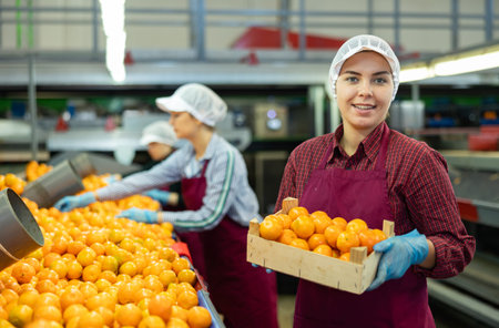 Smiling young female fruit warehouse worker with box of mandarins