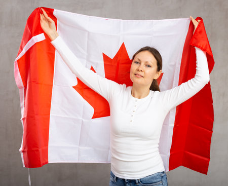 Positive patriotic middle-aged woman holding state flag of Canada against gray wall background indoorsの写真素材