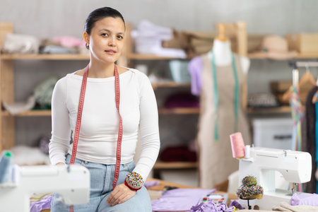 Stylish experienced young asian female dressmaker posing near work place with sewing machine and...