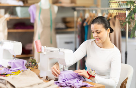 Positive young female dressmaker working with sewing-machine in tailors workshopの写真素材
