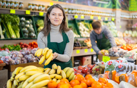 Young female seller holding bananas standing by counter in grocery marketの写真素材