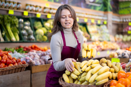 Positive female seller in apron displaying assortment of bananas at supermarketの写真素材