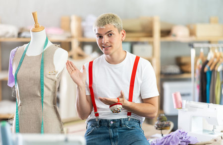 Stylish young male dressmaker posing near work place with sewing machine and tools, welcoming...