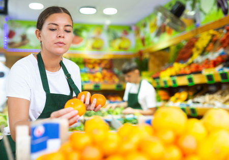 Smiling female seller standing at counter and lay out tangerines in grocery supermarketの写真素材