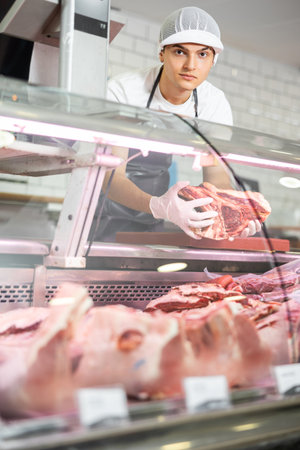 Positive young salesman demonstrating piece of meat in butcher shopの写真素材