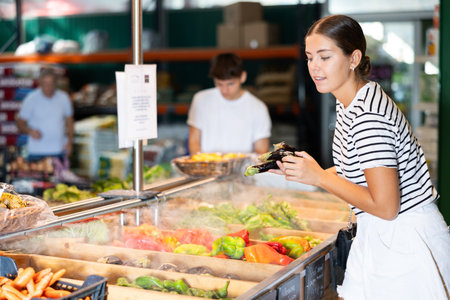 Smiling young female shopper choosing ripe eggplants in garden produce storeの写真素材