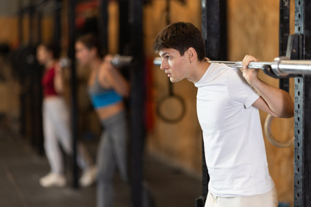 Slender guy trains muscles of arms and back using barbell and differentiated weighting in gym