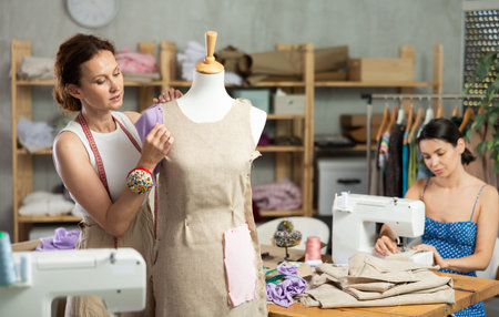 Female tailor sews new dress on mannequin in a sewing studio. Other woman tailor is sewing on sewing...