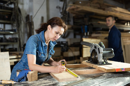 Woman employee of carpentry workshop makes measurements of wooden board, counts off set parameters