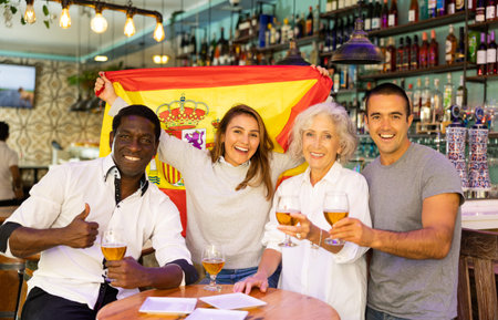 Excited diverse soccer supporters with flag of Spain celebrating victory with pint of beer in the...