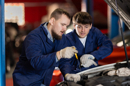 Two auto mechanics checking engine oil level under the hood of car together in garage