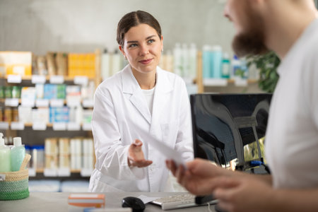 Male customer holding prescription searches for medicine in pharmacy. Female pharmacist offers to help find information on computerの写真素材