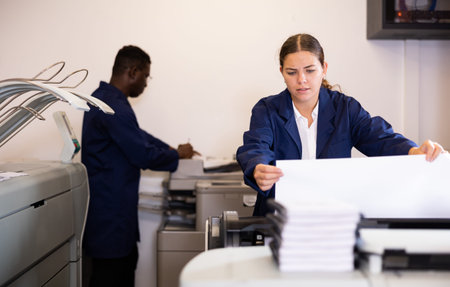 Serious young woman in uniform loading large format paper in a plotter in the print shop