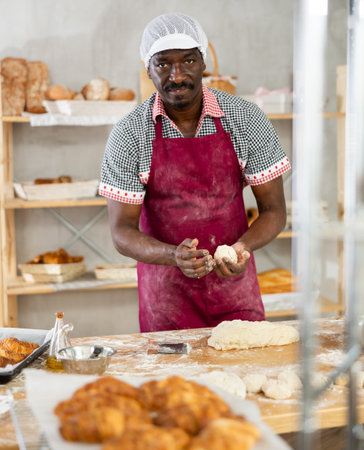 Confident male baker stands at his work bench, kneading and shaping dough to make bread, croissants...
