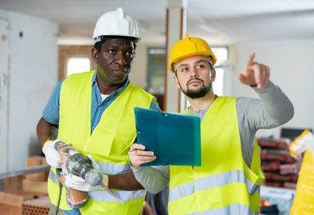 Two builders in with a hammer drill discuss work issues, making notesの写真素材