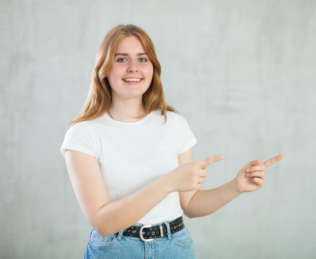 Portrait of positive teen girl in casual wear smiling at camera while pointing to right against concrete gray wall backgroundの写真素材