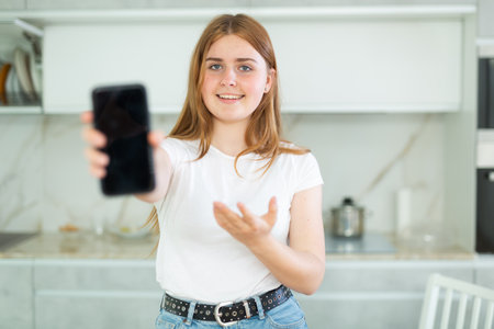 Smiling teenage girl presenting smartphone with blank black screen with copy space in modern cuisine. Free copy space for your design or advertisementの写真素材